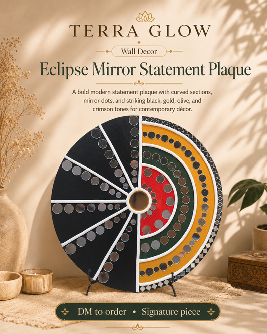 Terra Glow Eclipse Mirror Statement Plaque displayed on a styled tabletop with warm lighting and dried botanical props. The circular wall decor plaque features black, gold, red, green, and white mirrored sections with text above reading 'TERRA GLOW Wall Decor Eclipse Mirror Statement Plaque' and a 'DM to order • Signature piece' banner below.